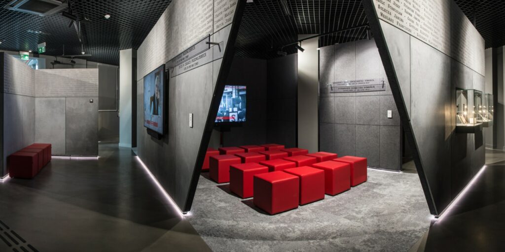 Fotografia przedstawia fragment przestrzeni wystawy znajdującej się w muzeum. Widoczne są szare ściany labiryntu, na których wiszą monitory z relacjami świadków historii. Przed monitorami ustawione są czerwone, kwadratowe pufy dla zwiedzających.