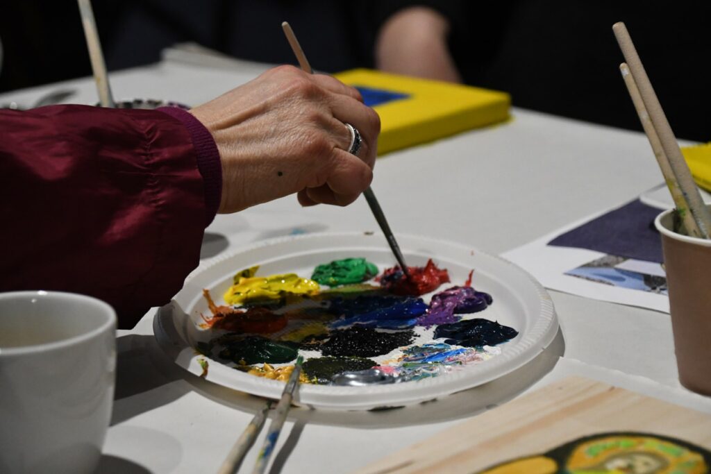 Zdjęcie przedstawia dłoń trzymającą pędzel nabierającą farby z palety znajdującej się na stoliku. Obok kubek z pędzlami. Zdjęcie wykonane w trakcie warsztatu pt.:"Anioł stróż z brzeszczańskiej apteki".