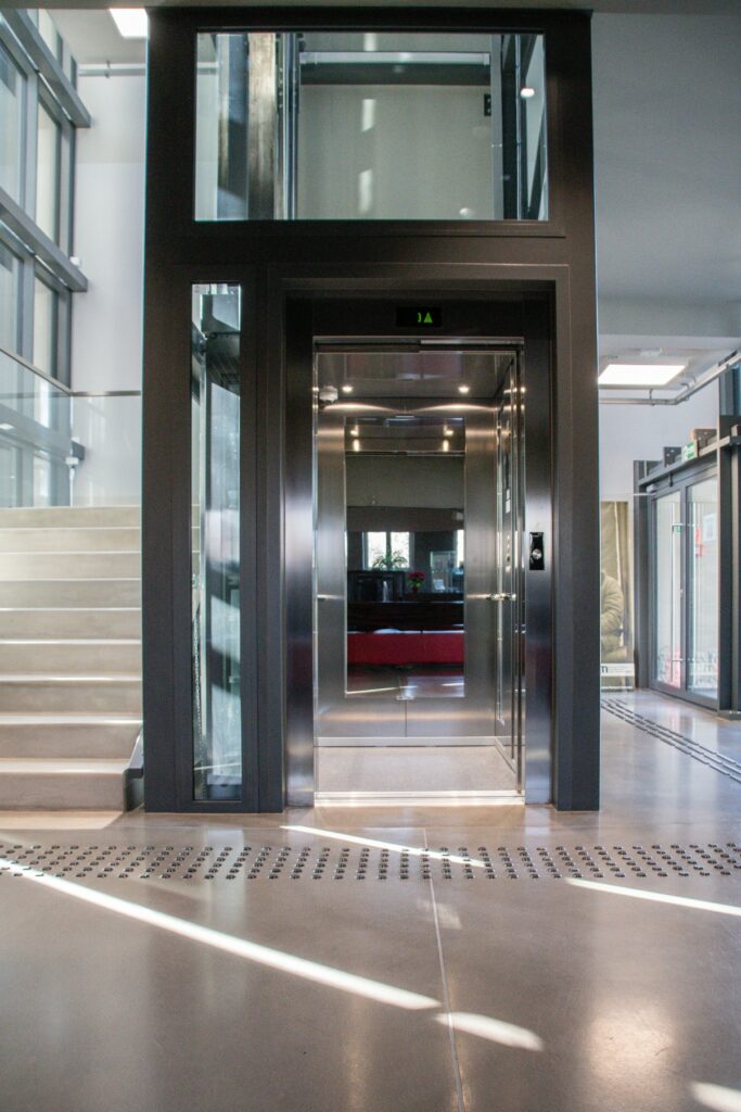 Fotografia przedstawia przeszkloną windę znajdującą się w budynku muzeum. Z lewej strony widoczne betonowe schody prowadzące na I piętro. Kolorystyka przestrzeni utrzymana w odcieniach szarości. Na podłodze, przed wejściem do windy oraz z prawej strony widoczne pola uwagi dla osób z niepełnosprawnością wzroku.