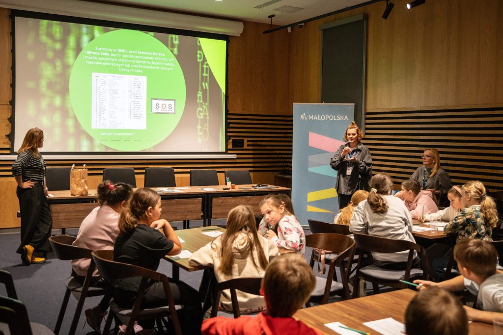 Sala koferencyjna. Przy stolikach siedzą dzieci. Na sali znajdują się trzy edukatorki prowadzące zajęcia. Na ekranie widoczny slajd z informacją o jednym z systemów szyfrowania.