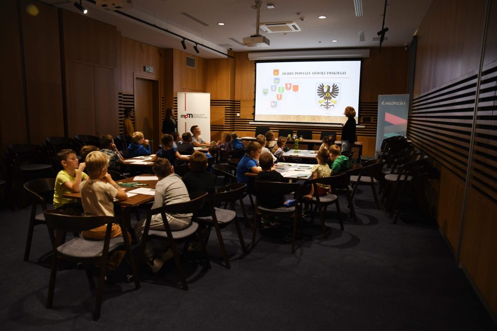 Przyciemniona sala koferencyjna. Przy stolikach siedzą dzieci. Na sali znajdują się trzy edukatorki prowadzące zajęcia. Na ekranie widoczny slajd z herbami z powiatu oświęcimskiego.