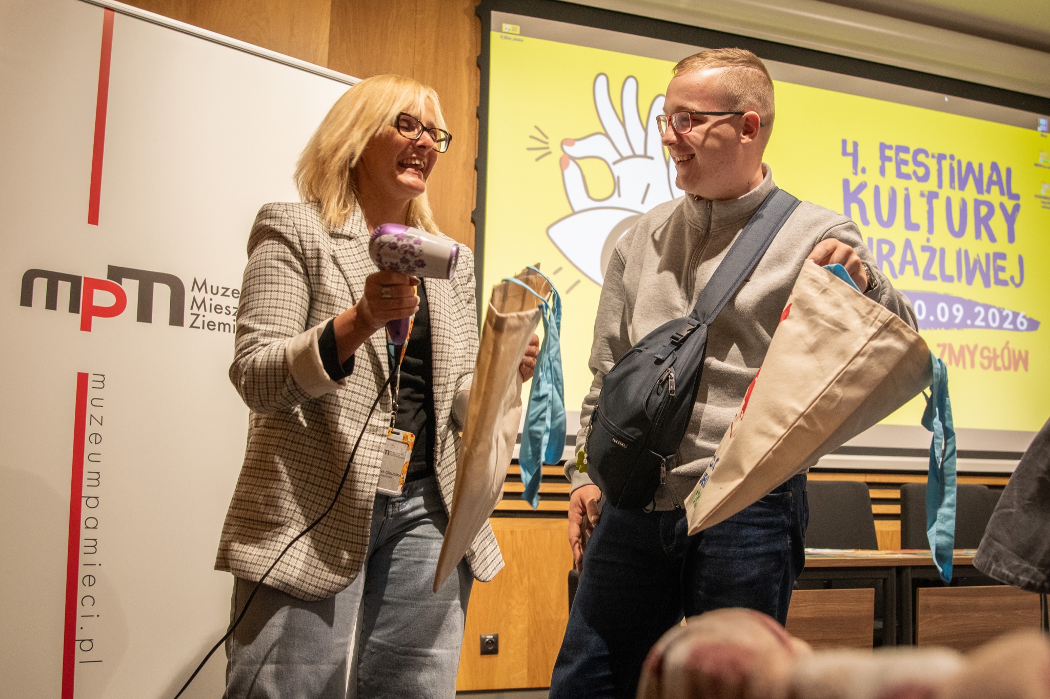 Dwie uśmiechnięte osoby tworzą projekt plastyczny. W tle banery festiwalu Kultury Wrażliwej i Muzeum Pamieci
