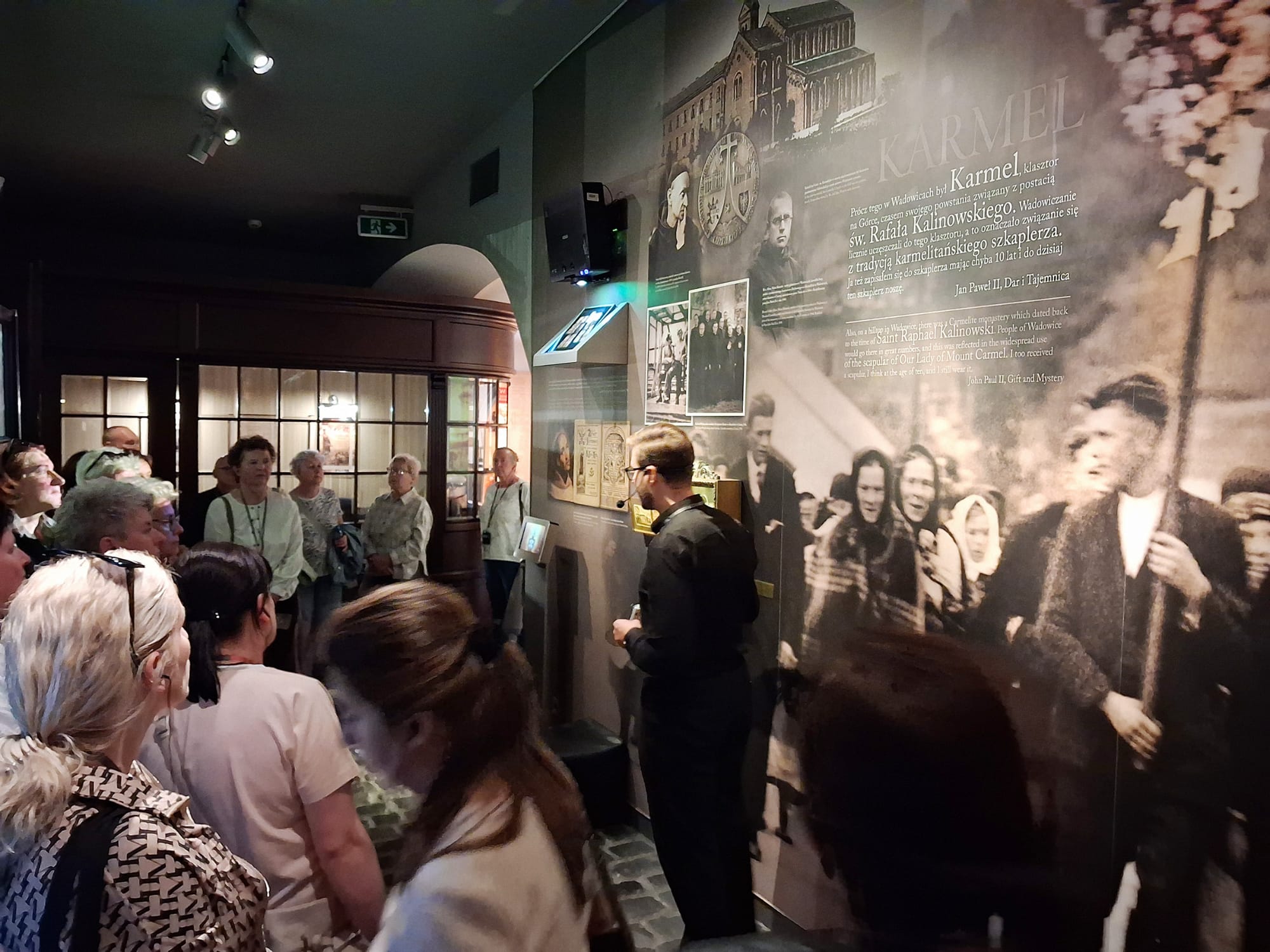 Zdjęcie wewnątrz budynku. W środku stoi przewodnik, który oprowadza ludzi po muzeum.