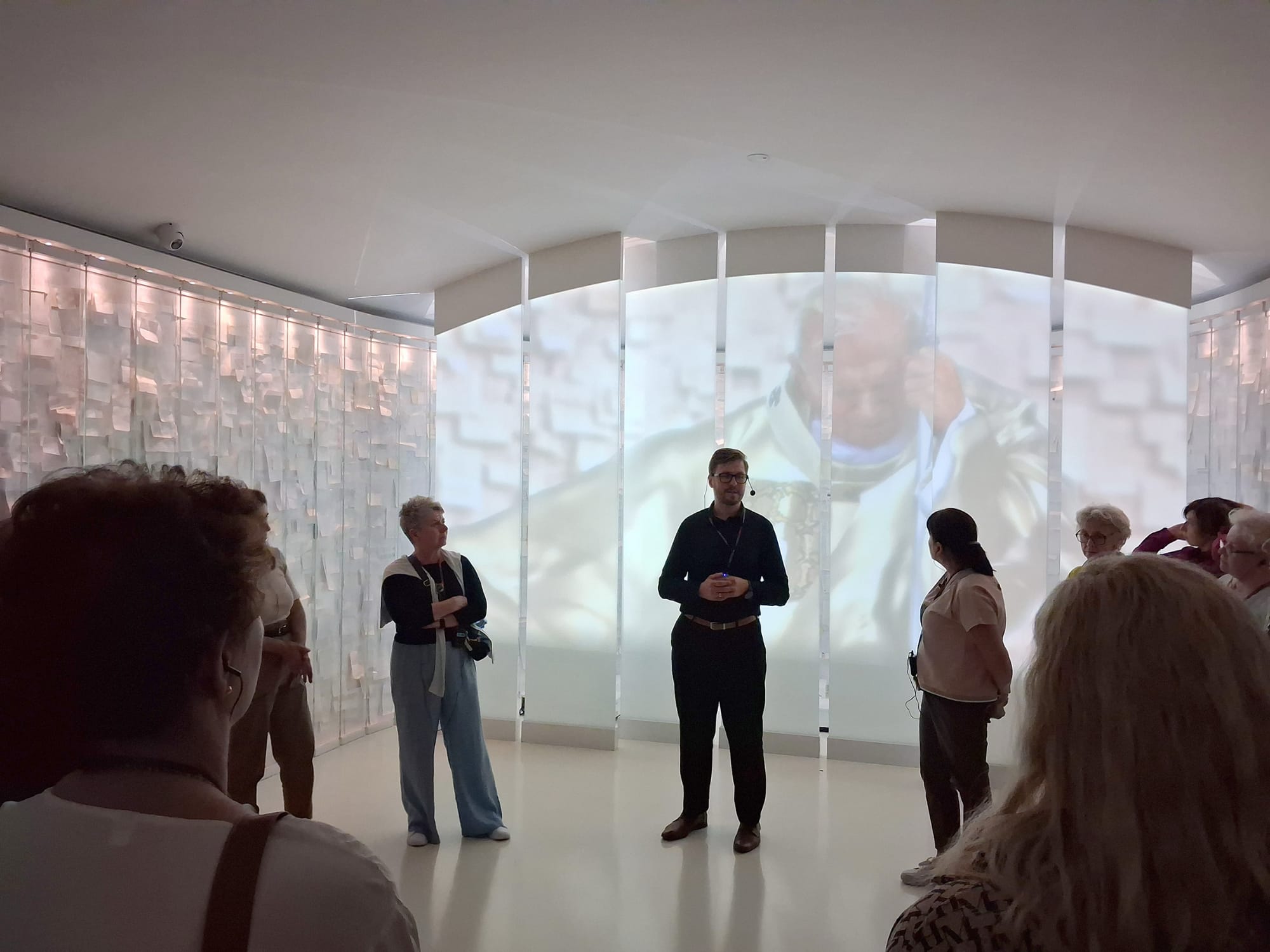 Mężczyzna będący przewodnikiem po muzeum, stoi na środku kadru. W tle zdjęcie Jana Pawła II.