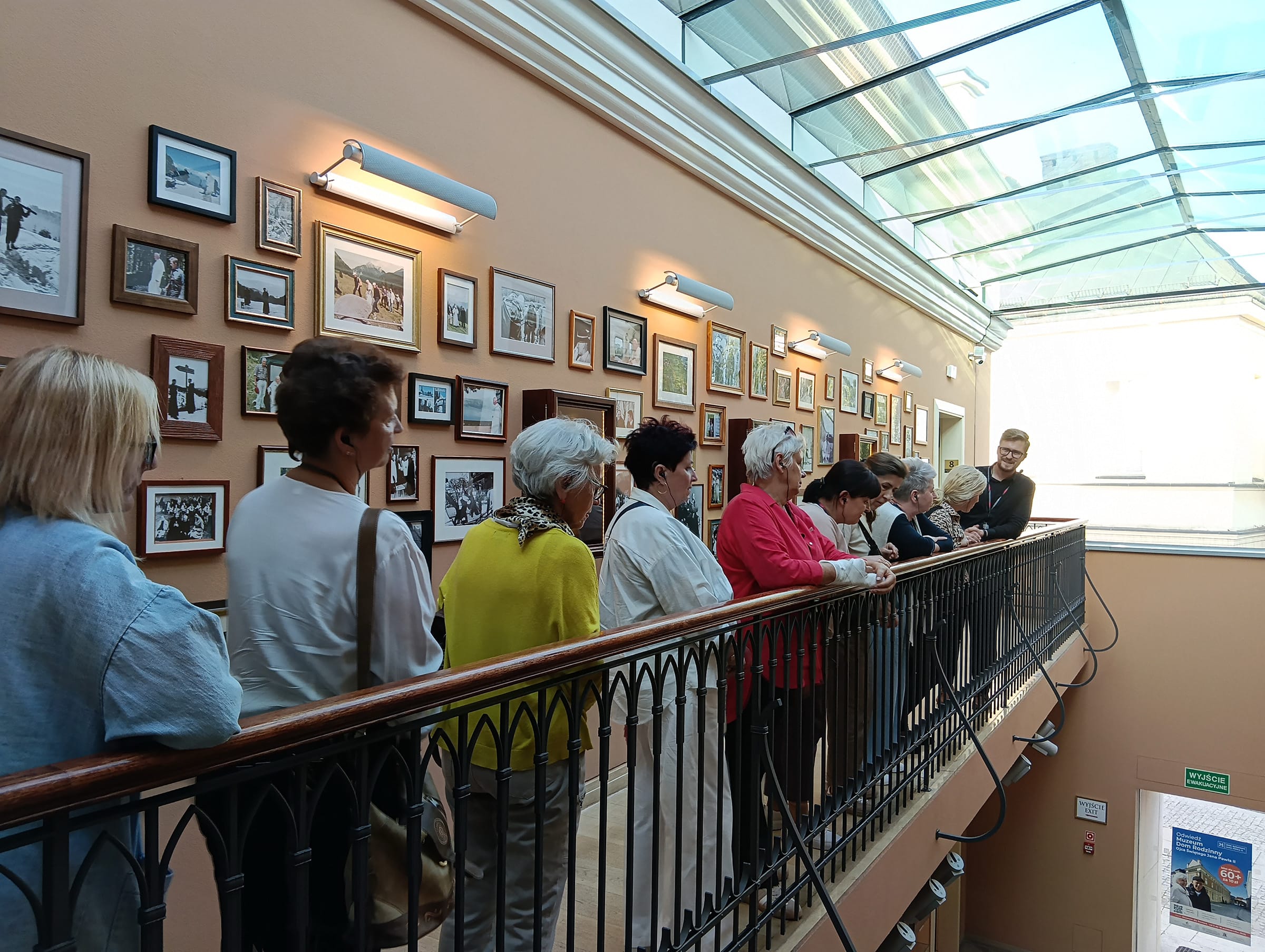 Grupa osób stoi na balkonie wewnątrz muzeum. W tle ściana z obrazkami w ramkach.