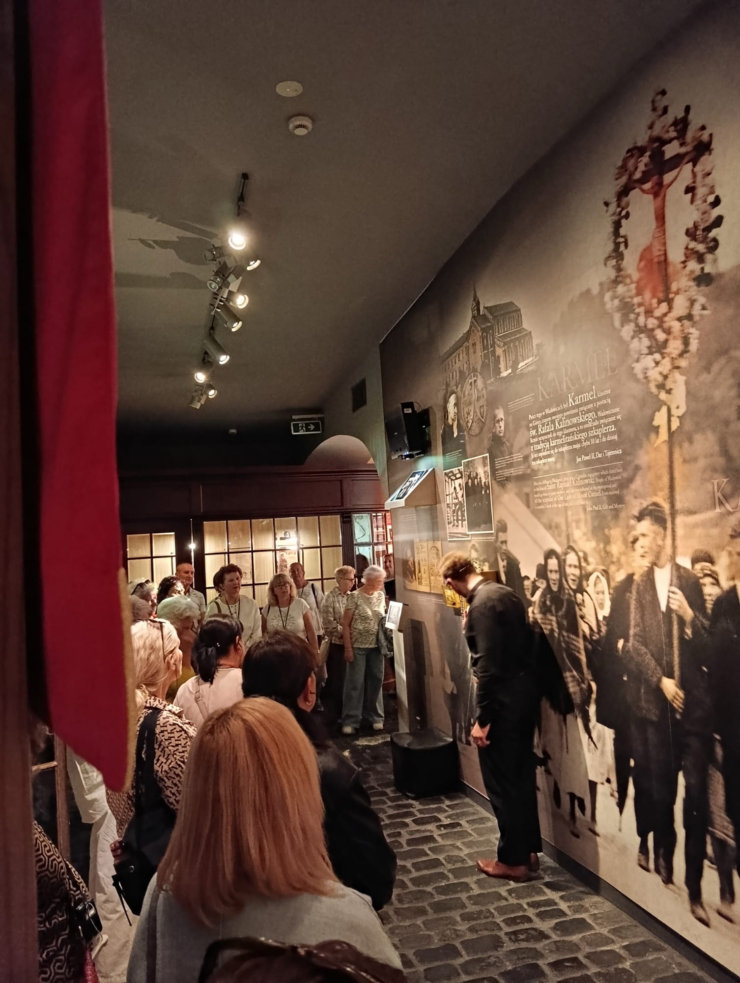 Grupa osób jest oprowadzana po muzeum. Na środku stoi przewodnik.