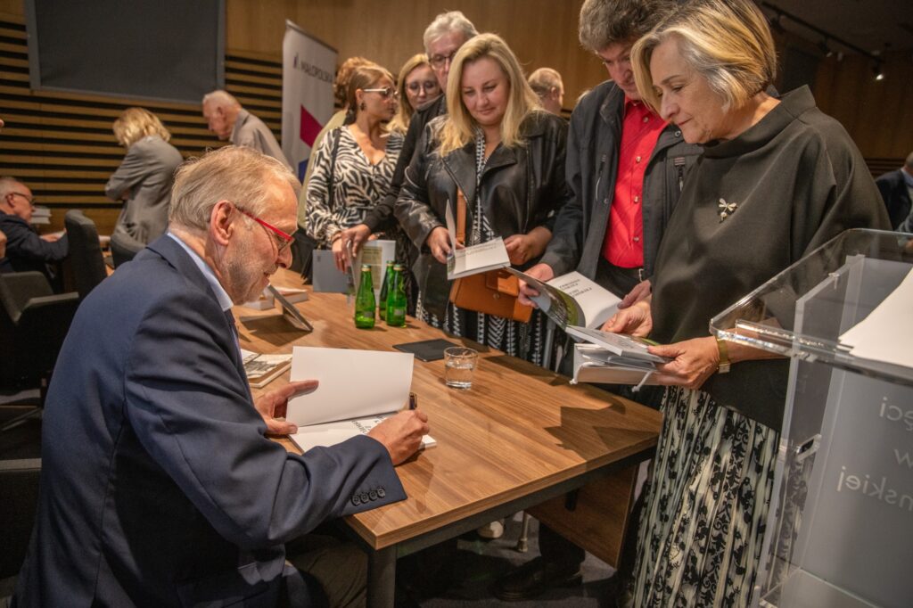 Mężczyzna siedzi za biurkiem i podpisuje książki. Przed biurkiem kolejka osób czekająca na podpis.