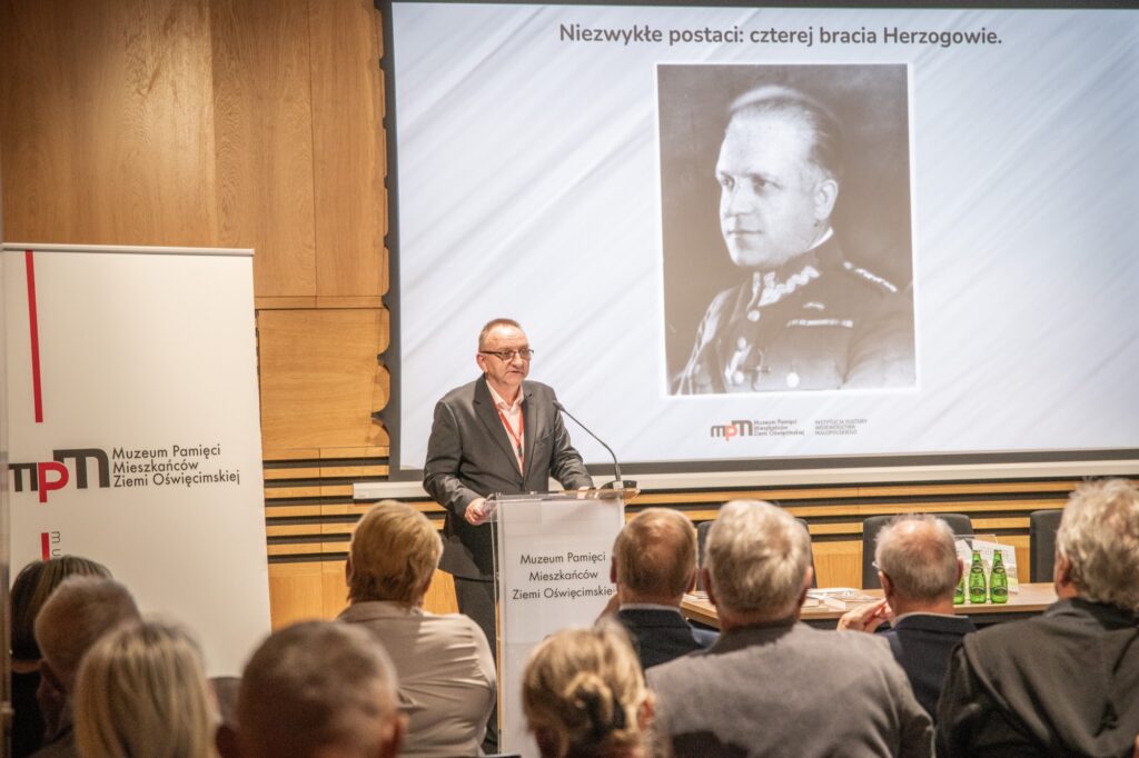 Mężczyzna na mównicy. Przemawia do osób zebranych w sali.