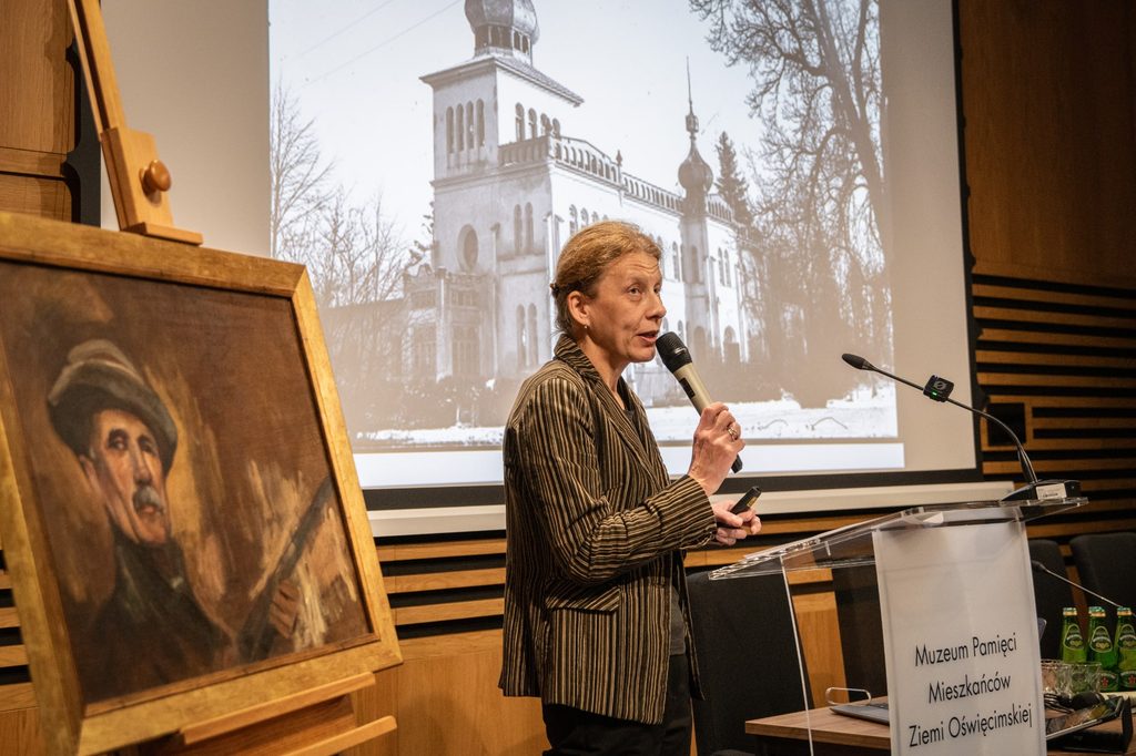 Kobieta trzyma mikrofon. Obok niej widać obraz. 
